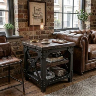 Industriální odkládací stolek ve stylu steampunk s nýtovaným černým ocelovým rámem a tmavou dřevěnou deskou vedle kožené pohovky Chesterfield v loftovém obýváku